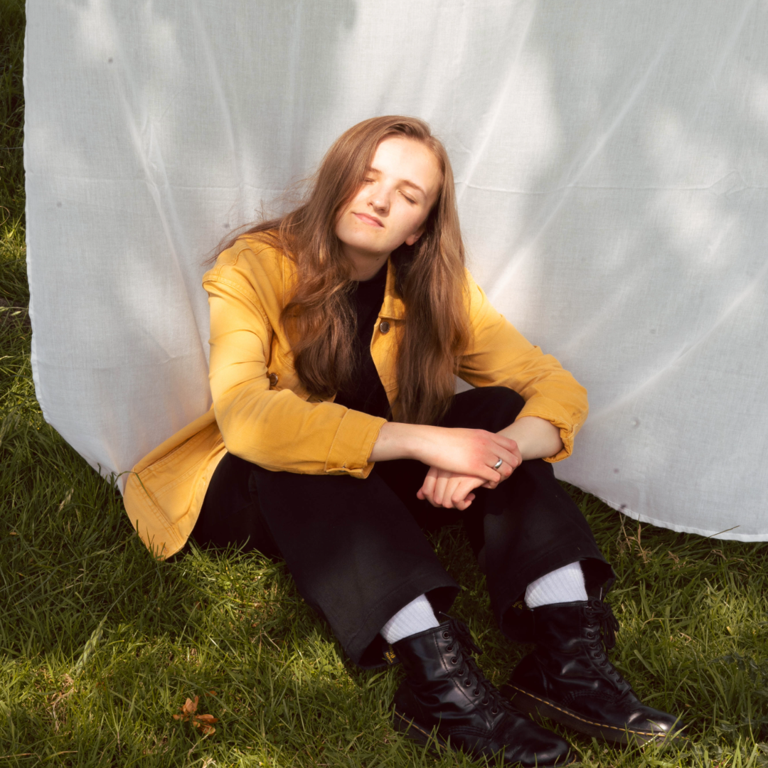 Evie Loose sitting on grass in a yellow jacket in front of a white sheet